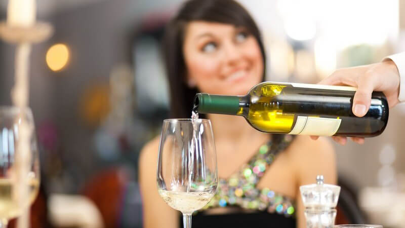 Kellner im Restaurant schenkt einer Dame ein Glas Weißwein ein, sie blickt freundlich zu ihm hoch