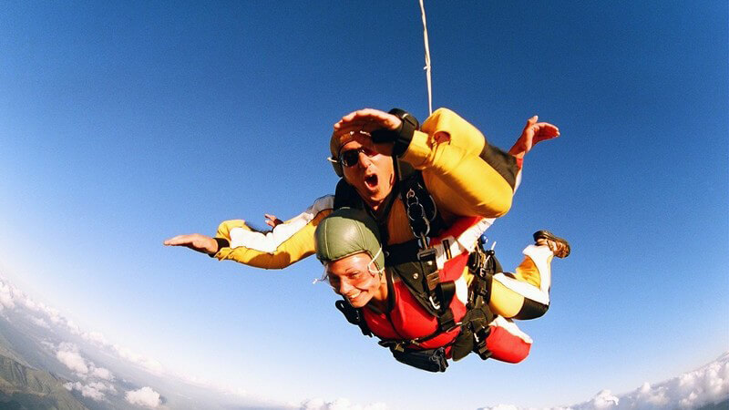 Tandem Fallschirmsprung unter blauem Himmel, im Hintergrund Bergpanorama