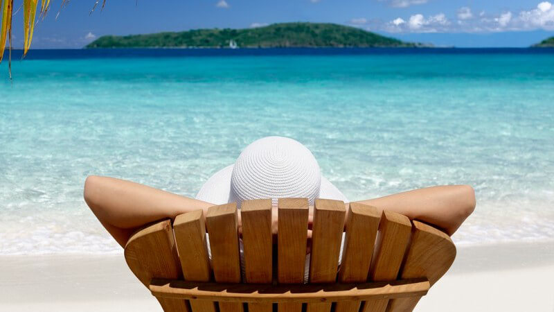 Urlauber mit Strohhut auf Sonnenliege unter Palmen mit Aussicht auf das Meer