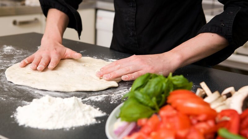 Pizzateig wird in Küche gemacht, daneben frische Tomaten