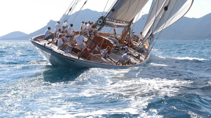 Segelschiff fährt auf dem Meer, Besatzung in weiß, im Hintergrund Berge