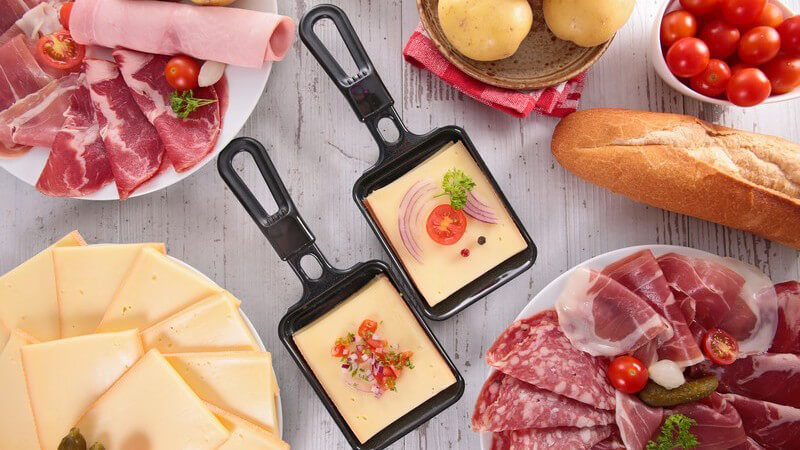 Zwei gefüllte Pfännchen und diverse Zutaten (Käse, Wurst, Kartoffeln) für Raclette