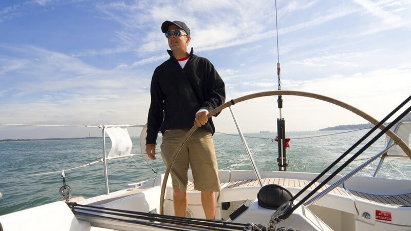 Mann mit Kappe und Sonnenbrille am Lenkrad eines Segelboots