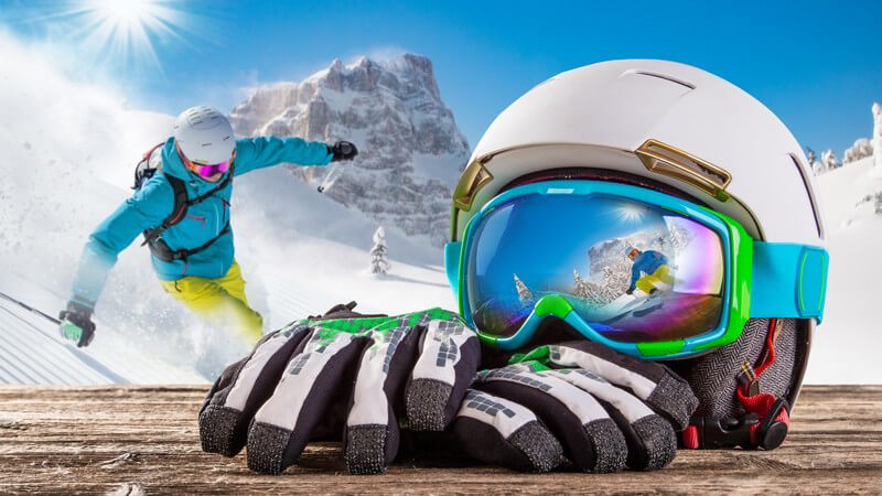 Weißer Skihelm und Skihandschuhe liegen auf einem Holztisch in der Sonne, im Hintergrund die Skipiste und ein Skifahrer