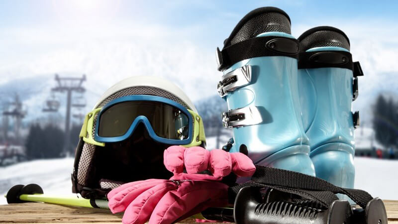 Skihelm, Skischuhe, Stöcke und Handschuhe auf rustikalem Holztisch, im Hintergrund ein Skigebiet