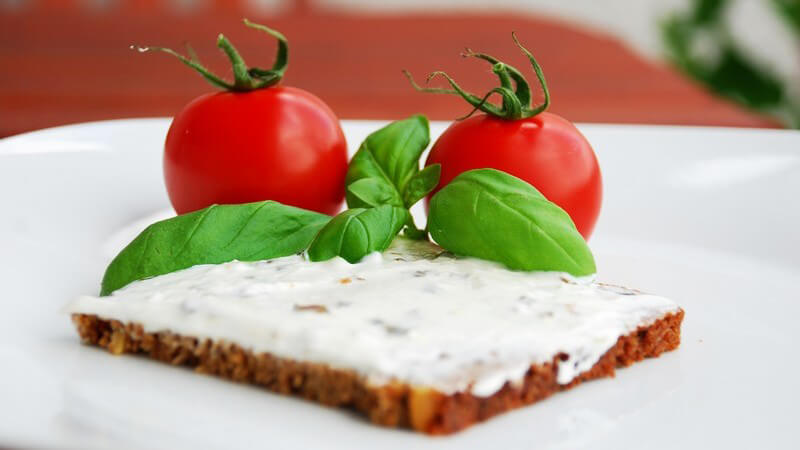 Scheibe Brot mit Frischkäse, dahinter frische Tomaten und Basilikum