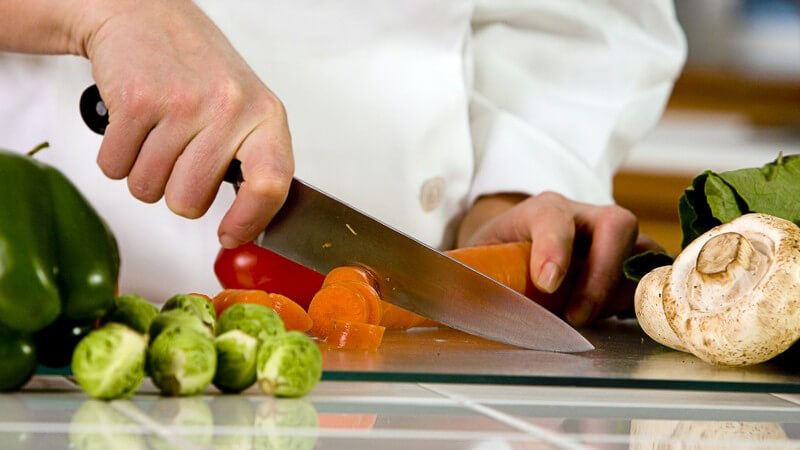 Nahaufnahme Koch schneidet Karotten, daneben verschiedenes frisches Gemüse