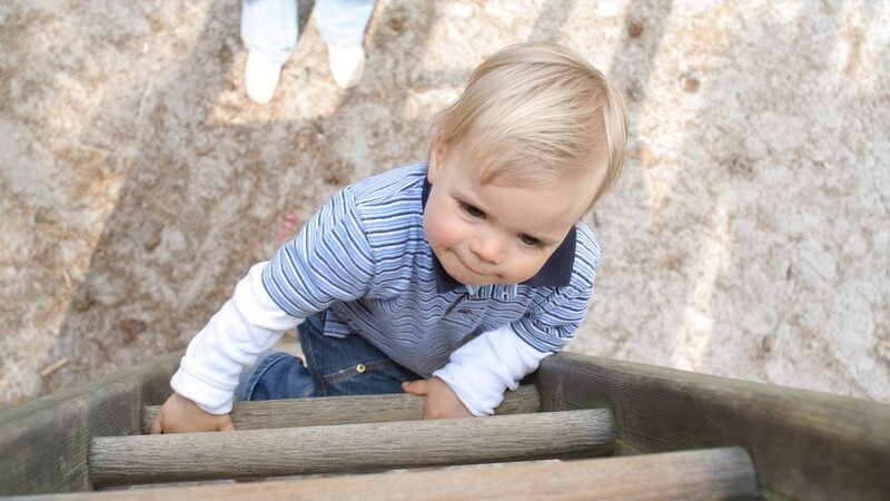 Aufnahme von oben, Kleinkind klettert Holzleiter hinauf, unten steht Mutter