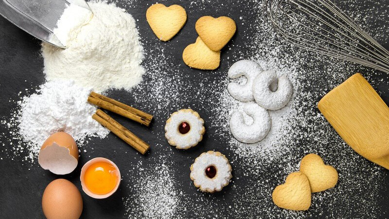 Backzutaten wie Mehl und Eier sowie fertige Weihnachtsplätzchen auf dunklem Untergrund