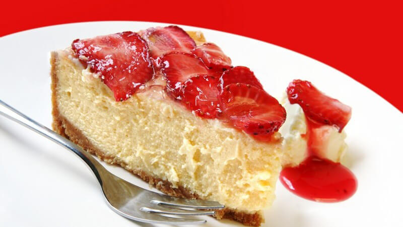 Erdbeerkuchen auf weißem Teller mit Gabel vor rotem Hintergrund, ein einzelnes Stück mit Sauce daneben