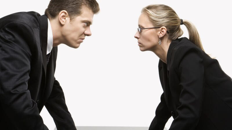 Frau und Mann mit bösem Gesichtsausdruck stehen sich gegenüber, Hände auf Tischplatte