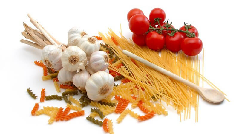 Auswahl an bunten Nudeln, darauf Knoblauch, Kochlöffel und frische Tomaten