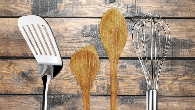 Pfannenwender, Rührbesen und zwei Holzkochlöffel vor Holzwand