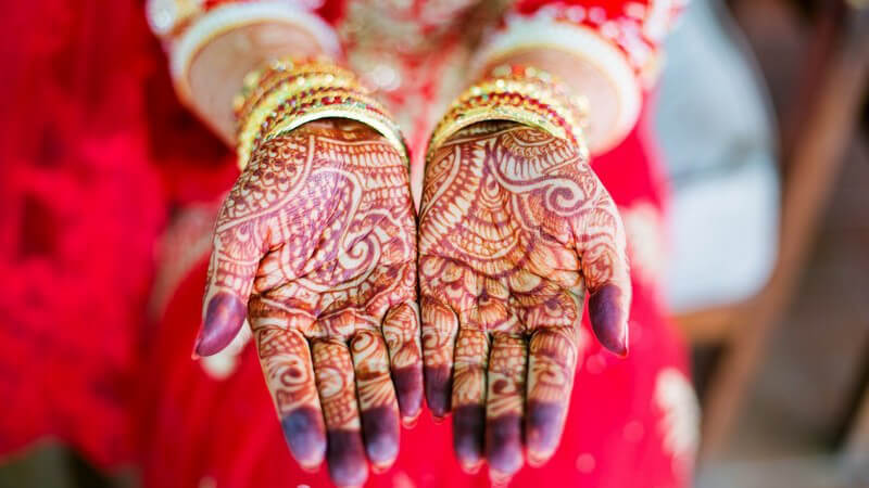 Indische Braut in rotem Kleid und mit buntem Armschmuck zeigt ihre Handinnenflächen mit lila Henna-Tattos