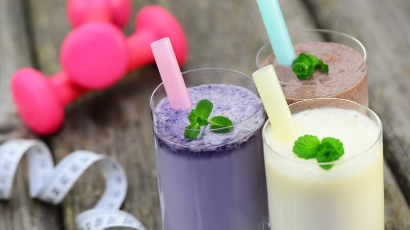 Drei unterschiedliche Milchshakes mit Strohhalm, daneben pinke Hanteln und ein Maßband