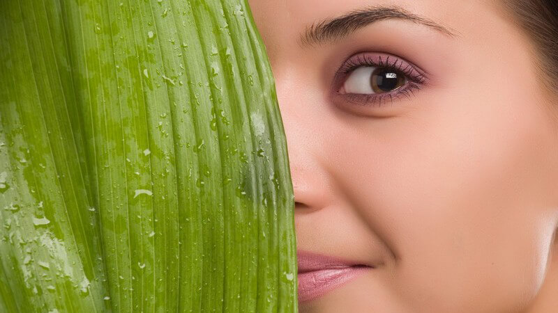 Junge, dunkelhaarige Frau versteckt ihr Gesicht halb hinter grünem Blatt