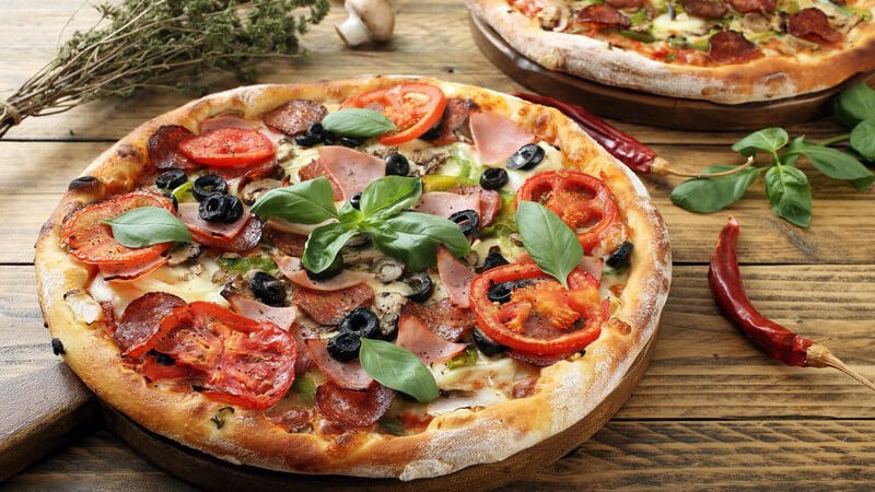 Frisch gebackene Pizza mit Tomaten, Basilikum, Oliven und Wurst auf rustikalem Holztisch