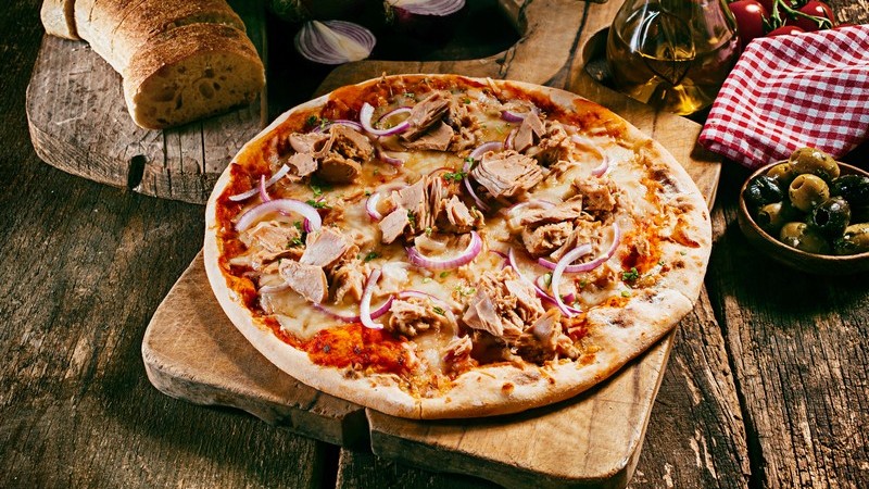 Thunfischpizza mit Zwiebeln auf Holzbrett, daneben Brot und Oliven