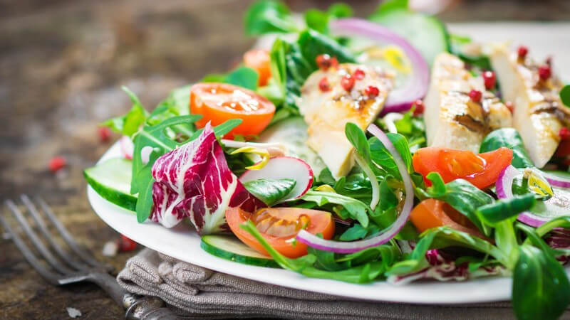 Bunter Salatteller mit Hähnchenbruststreifen auf einem grauen Tuch, daneben eine Gabel
