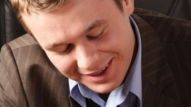 Junger Mann in Nadelstreifenanzug sitzt lächelnd auf Chefsessel oder Bürostuhl und hat die Augen niedergeschlagen