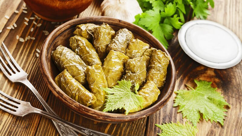 Gefüllte Weinblätter (Dolmas oder Dolmades) in einer braunen Schale neben Gabeln und Kräutern auf Holztisch