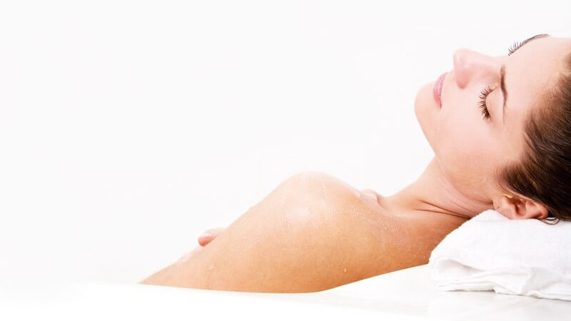 Dunkelhaarige Frau liegt entspannt in der Badewanne, den Kopf auf einem weißen Handtuch