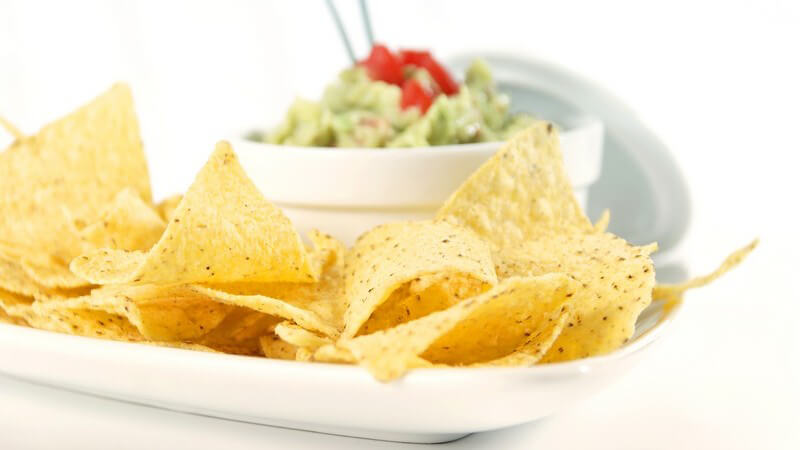 Eine Platte Tortillachips mit grün-rotenm Guacamole-Salat im Hintergrund, in dem Stäbchen stecken