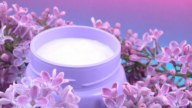 Bodylotion im lilanen Döschen, umgeben von frischem Flieder