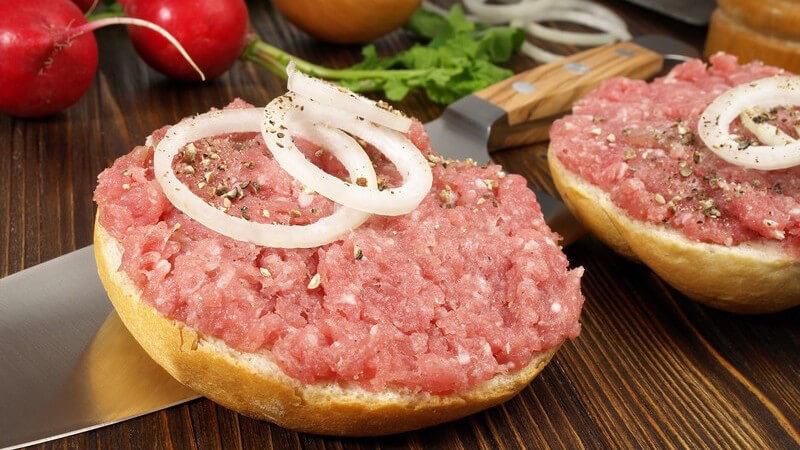 Zwei Hälften Schweinemettbrötchen mit Zwiebelringen, neben Kochmesser, Radieschen und Zwiebel