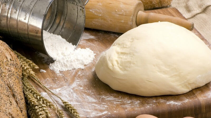 Roher Teig auf Holzbrett neben Mehl, Eiern, Nudelholz, Getreide