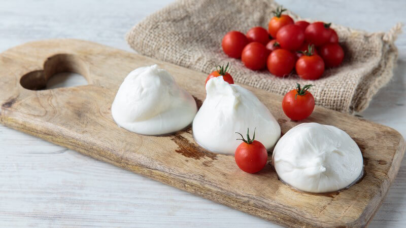 Drei Häufchen Burrata-Käse auf Holzbrett, daneben liegen Kirschtomaten auf Jutesack