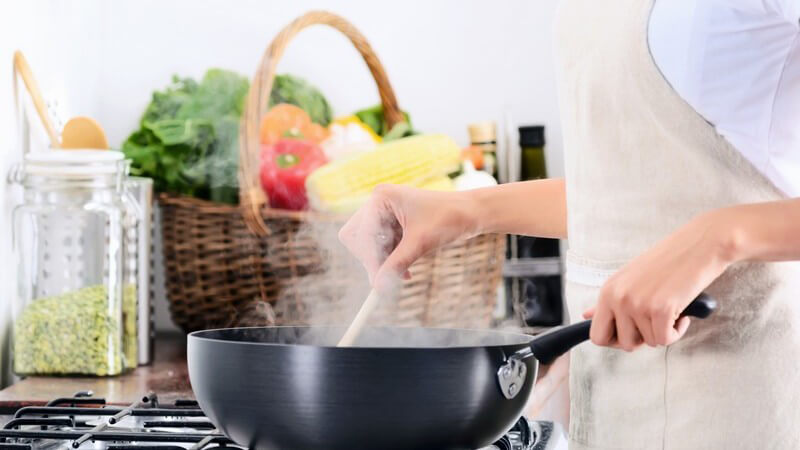 Frau mit Schürze steht am Gasherd und rührt Essen in einer hohen Pfanne um