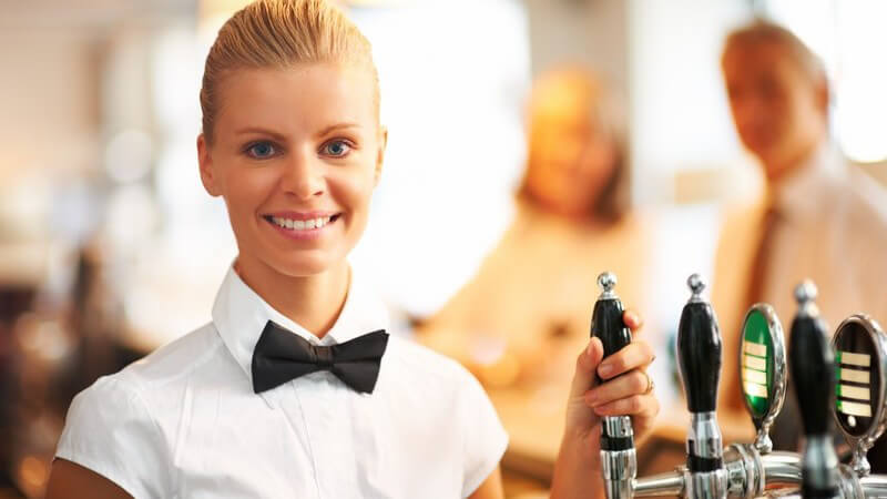 Junge Kellnerin mit Fliege vor Zapfanlage, lächelt in Kamera