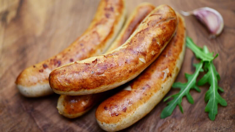 Vier gegrillte Rostbratwürste neben Rucolablättern und Knoblauch auf einem Holztisch