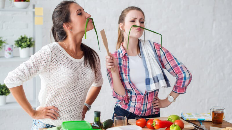 Zwei junge Frauen spielen beim gemeinsamen Kochen mit den Zutaten, benutzen Frühlingszwiebeln als Schnurrbart