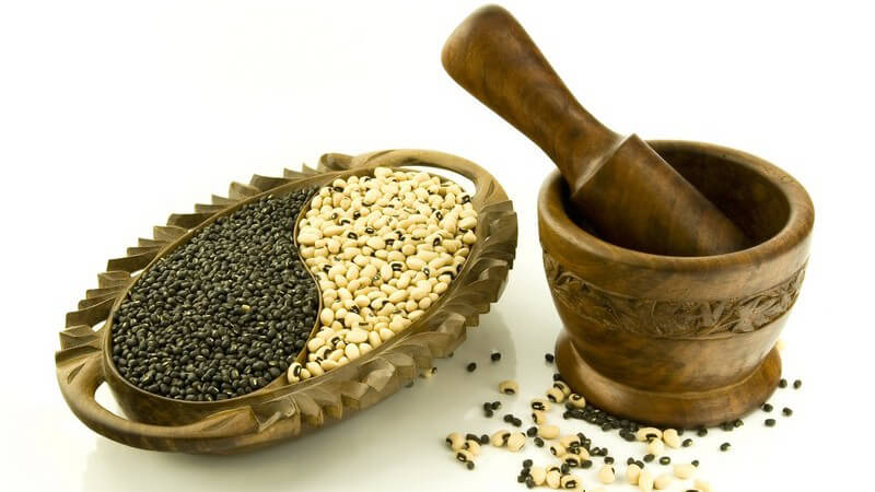 Ovales Holzgefäß gefüllt mit weißen und schwarzen Bohnen, daneben Holzmörser, beides mit Schnitzungen