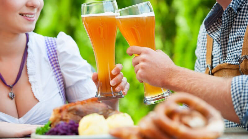 Paar in Trachtenkleidung (Dirndl, Lederhose) stößt mit Weizenbier an und isst zünftiges Gericht (Knödel)