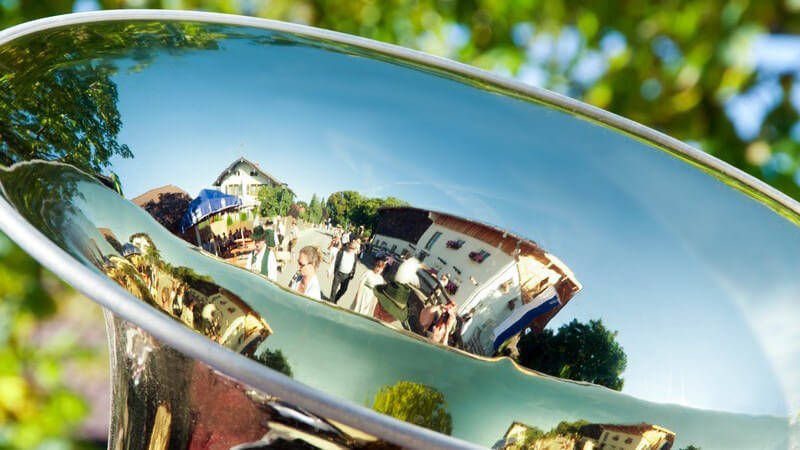 Nahaufnahme einer Tuba, in der sich bei strahlendem Sonnenschein Menschen und Häuser spiegeln