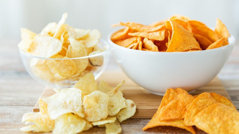 Kartoffelchips in Glasschüssel und Nachos in weißer Schüssel auf Holzbrett