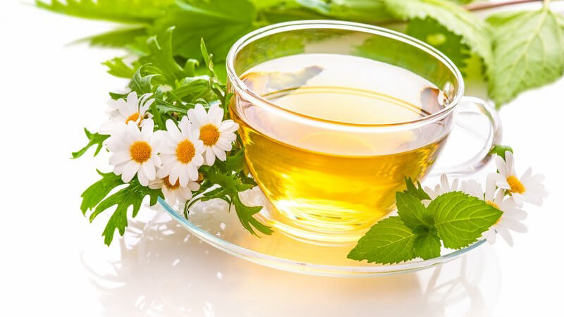 Glastasse mit gelbem Kräutertee, umgeben von Kamille und Minze