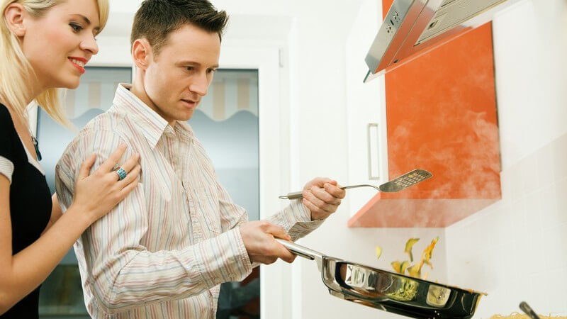 Paar steht vor dem Herd, Mann schwenkt Pfanne mit Essen und hält Pfannenwender