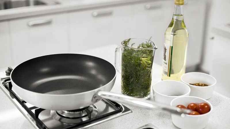 Pfanne auf Gasherd, daneben Kräuter und Gemüse
