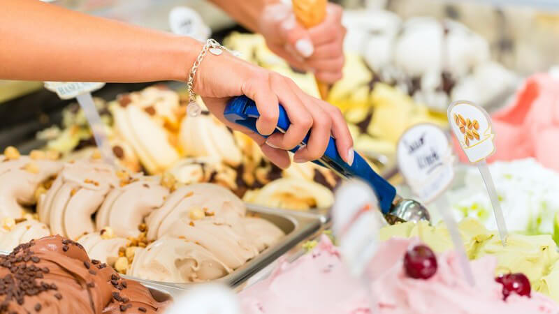 Blick auf das Eissortiment in einer Eisdiele, ein Hörnchen wird gerade mit Eiskugeln befüllt