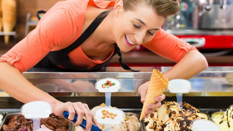 Eisverkäuferin mit Schürze füllt in einer Eisdiele ein Hörnchen mit Eiskugeln