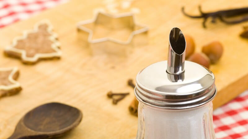 Zuckerstreuer vor einem Holzbrett mit Plätzchen, Ausstechern, Löffel und Zutaten