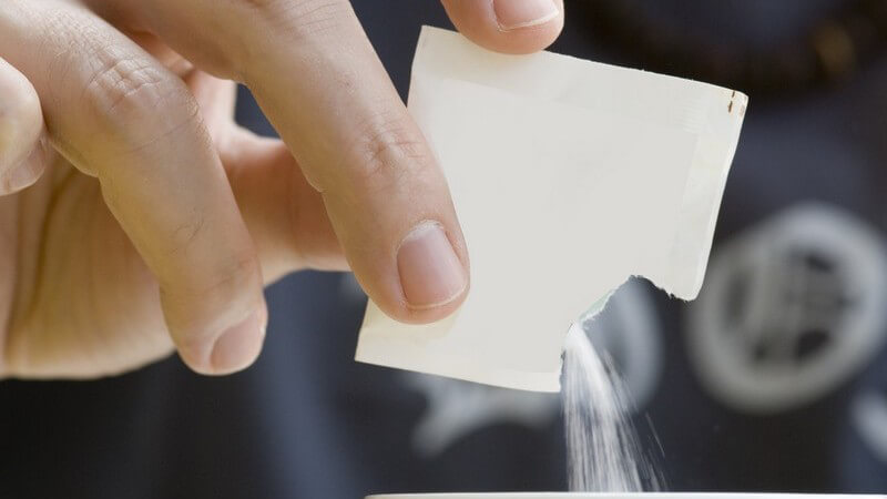Männerhand, die Zucker aus einem weißen Zuckertütchen in den Kaffee in der Kaffeetasse schüttet