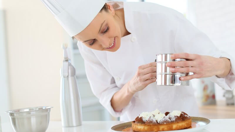 Köchin mit Kochmütze streut Puderzucker über einen Kuchen