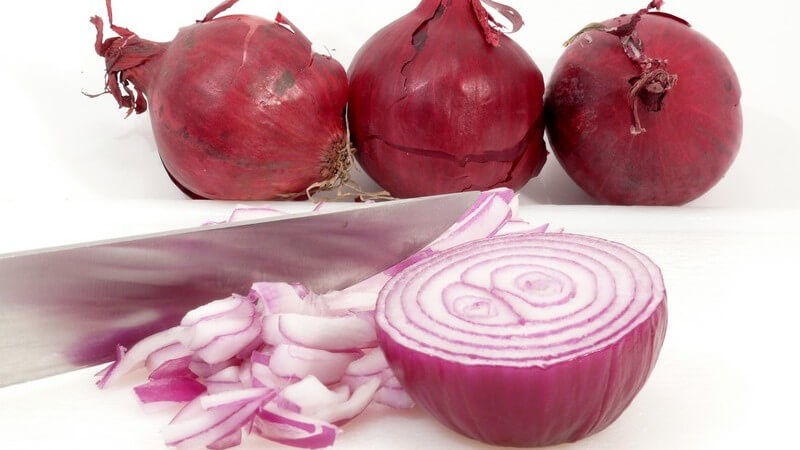 Drei rote Zwiebeln, davor aufgeschnittene Zwiebel neben Messer auf weißem Hintergrund