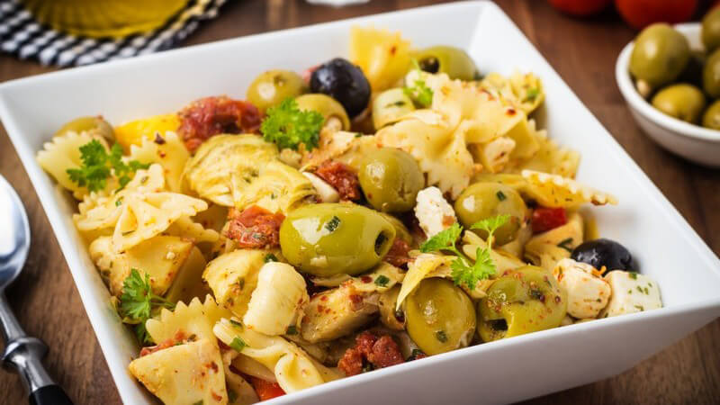 Mediterraner Nudelsalat mit Oliven und getrockneten Tomaten in einer weißen viereckigen Schüssel auf einem Holztisch