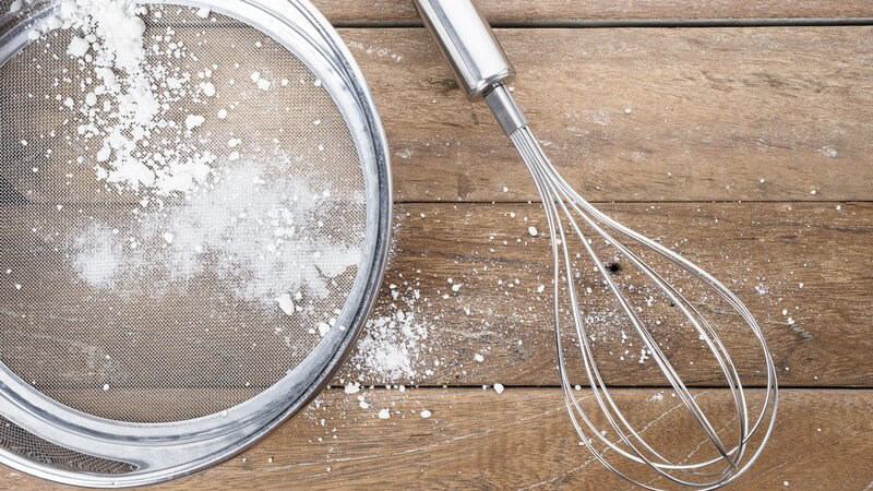 Feines Sieb mit Puderzucker neben Rührbesen auf Holztisch